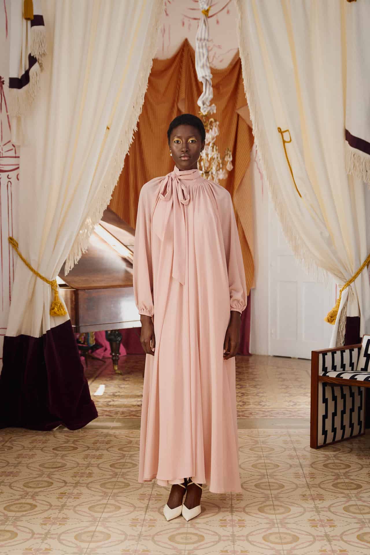 Mujer con vestido largo rosa empolvado de manga larga y lazo al cuello, en un entorno elegante con cortinas y piano de fondo. Look sofisticado ideal para madres o invitadas a una comunión.